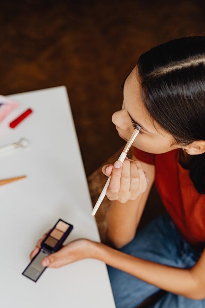 A young woman carefully applies eyeshadow using a brush, captured in a high-angle shot.